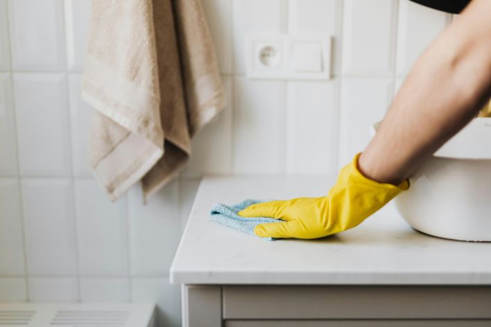 Close-up of a person wearing yellow gloves wiping a bathroom counter with a cloth.